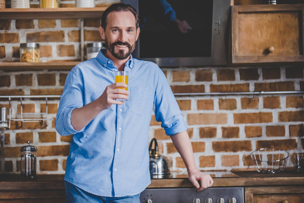 smiling mid adult man holding orabge juice at the kitchen - Foto, Imagem