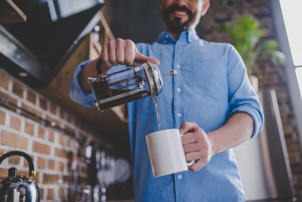 low angle view of happy man pouring coffee into the cup at morning in the kitchen, cropped image - Fotó, kép