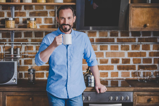 smiling mid adult man holding coffee cup and looking at the camera at the kitchen - 写真・画像