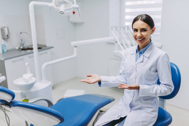 πορτρέτο του χαμογελαστό γυναίκα οδοντίατρος που δείχνει τη θέση της εργασίας στη σύγχρονη ιατρική κλινική - Φωτογραφία, εικόνα