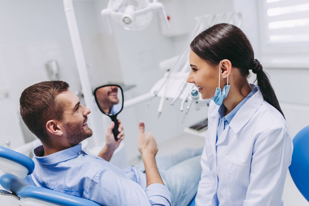 smiling male patient looking in the mirror cheking teeth after dental procedure in clinic - Fotoğraf, Görsel