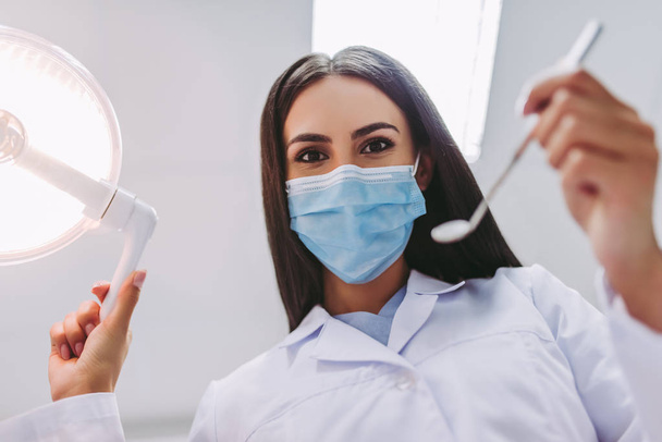 vista de bajo ángulo del dentista en máscara médica que sostiene herramientas dentales en la clínica dental moderna
 - Foto, imagen