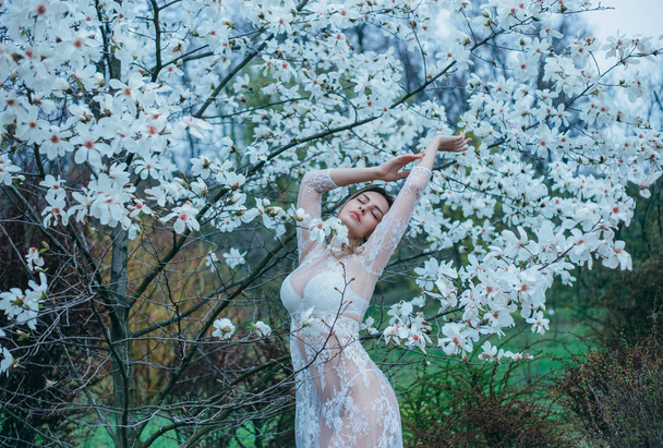 A girl with fair hair, in a transparent dressing-gown posing against a background of blooming, white magnolia. Spring, gentle portraits, as much as possible natural with minimal processing - Foto, afbeelding