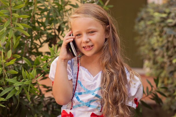 Adorable girl in princess dress and with long curly hair using smartphone and speaking on street in summertime - Foto, afbeelding