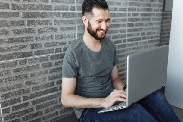 Portrait of a beautiful stylish guy, a hipster, running on a laptop, wearing a gray empty t-shirt, standing on a brick gray wall background. - Foto, imagen