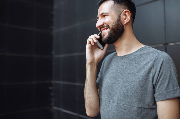 Portrait of a handsome stylish guy, hipster, talking on the phone, wearing a gray empty t-shirt, standing on a black wall background. - Fotoğraf, Görsel