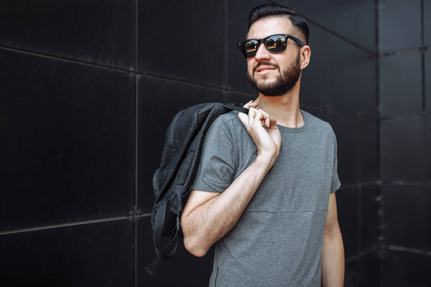 Portrait of a beautiful stylish guy, hipster with glasses, dressed in a gray empty t-shirt, standing on a black wall background. - Фото, изображение
