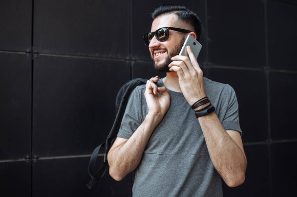 Portrait of a beautiful stylish guy, hipster with glasses, talking on the phone, dressed in a gray empty t-shirt, standing on a black wall background. - Photo, image