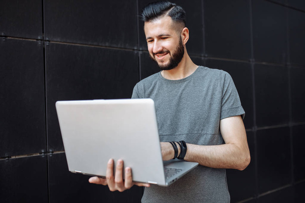 Portrait of handsome stylish guy, the hipster, working on laptop, wearing a gray blank t-shirt standing against a black wall. - 写真・画像
