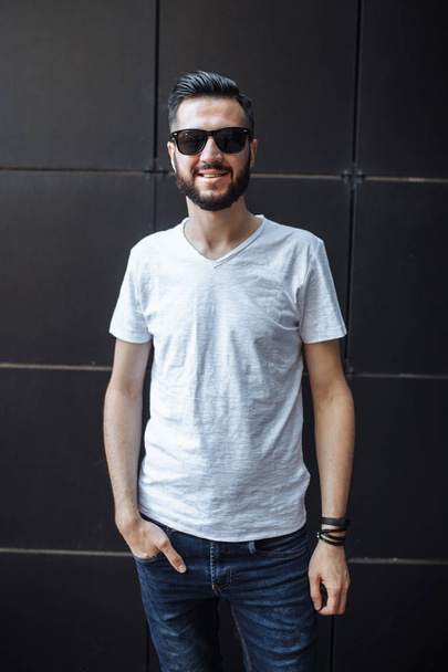 Photo of a beautiful stylish guy, a hipster with glasses, dressed in a white empty t-shirt, standing on a black wall background. . - Φωτογραφία, εικόνα