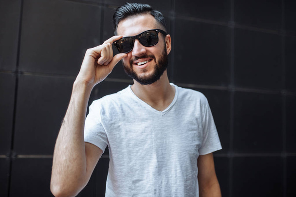 Photo of a beautiful stylish guy, a hipster with glasses, dressed in a white empty t-shirt, standing on a black wall background. . - Фото, изображение