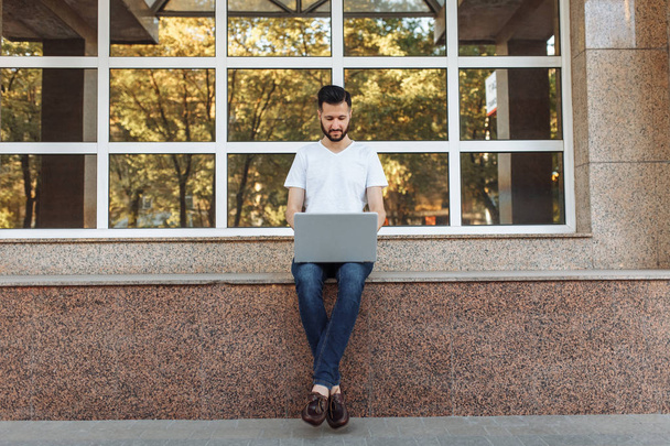 Portrait of a beautiful stylish guy dressed in a white t-shirt, who sits on a parapet, works with a laptop, on the street against a glass building, - 写真・画像
