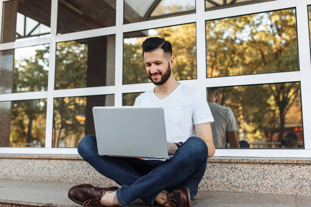Portrait of a beautiful stylish guy dressed in a white t-shirt, who sits on a parapet, works with a laptop, on the street against a glass building, - Zdjęcie, obraz