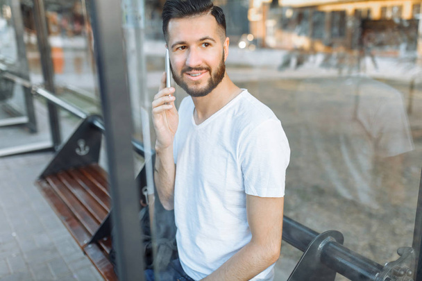 photos of a beautiful stylish Guy, in a blank white t-shirt, a hipster talking on the phone, and sitting at the bus stop waiting for transport, - Valokuva, kuva