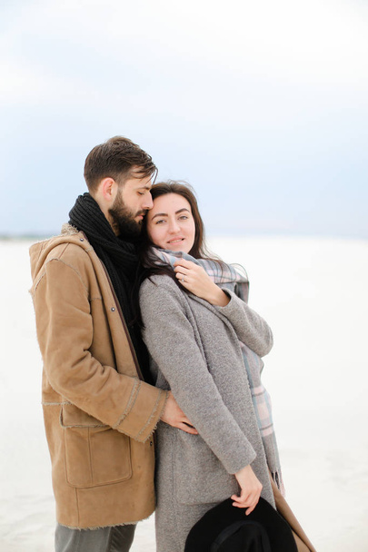 Щасливий молодий чоловік з бородою обіймає жінку в сірому пальто та шарфі, білий зимовий однотонний фон
. - Фото, зображення