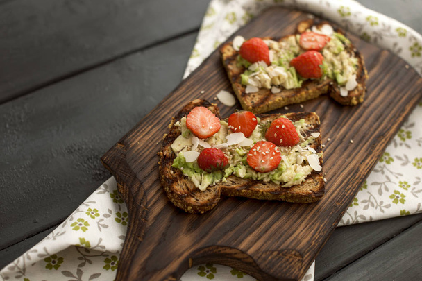 Toasts with avocado and strawberries, on grain bread for breakfast. White napkin with spikes and a black wooden background. copy space - Фото, изображение