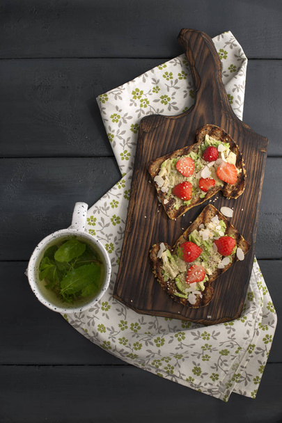 tea with fresh mint in a white mug on a napkin and a black wooden background. Healthy breakfast toast with avocado and strawberries. Copy space, flat lay - Фото, изображение