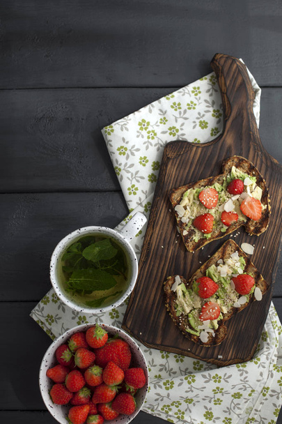 tea with fresh mint in a white mug on a napkin and a black wooden background. Healthy breakfast toast with avocado and strawberries. Copy space, flat lay - Valokuva, kuva
