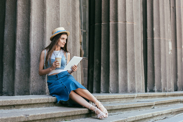 beautiful smiling young woman in hat holding paper cup and digital tablet while sitting on stairs and looking away - Photo, Image