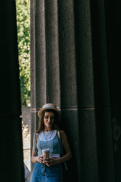 beautiful smiling young woman in hat holding coffee to go and standing between columns - 写真・画像