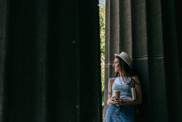 красива усміхнена молода жінка з книгою і кавою, щоб йти стояти між колонками і дивитися
 - Фото, зображення