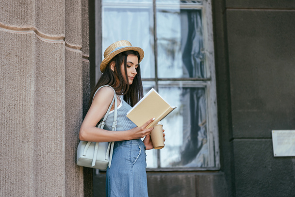side view of attractive girl in hat holding coffee to go and reading book near column - Foto, imagen