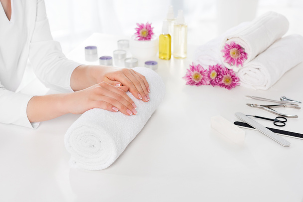 partial view of woman holding hands at table with towels, flowers, candles, aroma oil bottles and instruments for manicure in beauty salon - Photo, Image