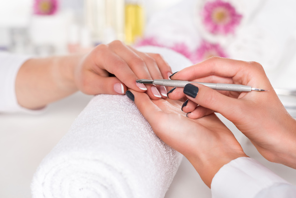 close up image of  woman receiving manicure by beautician with cuticle pusher at table with flowers and towels in beauty salon  - Foto, immagini