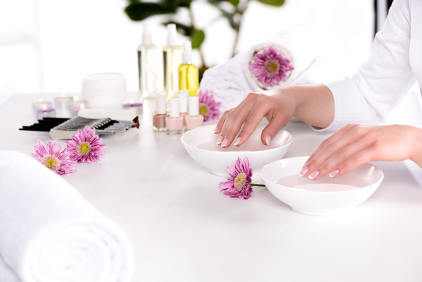 cropped image of woman receiving bath for nails at table with flowers, towels, candles, aroma oil bottles, nail polishes, cream container and tools for manicure in beauty salon  - Photo, Image