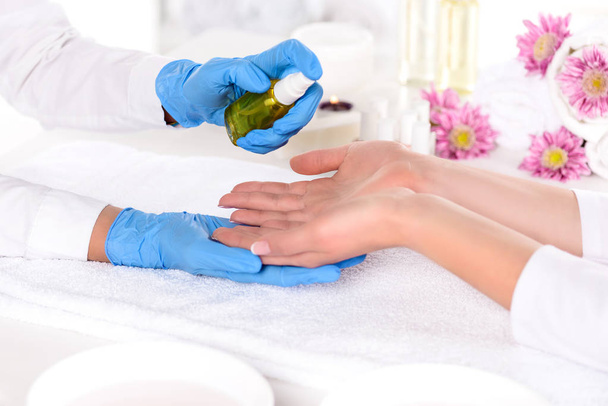 cropped image of manicurist in latex gloves spraying aroma oil on hands of woman in beauty salon - Фото, изображение