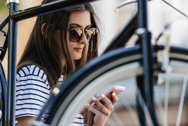 portrait of young woman using smartphone while sitting near retro bicycle on street - Фото, изображение