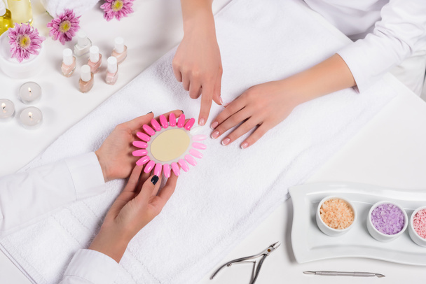 cropped image of manicurist holding samples of nail varnishes while woman pointing by finger at table with aroma oil bottles, candles, towels, flowers, nail polishes, sea salt and nail clippers - Photo, Image