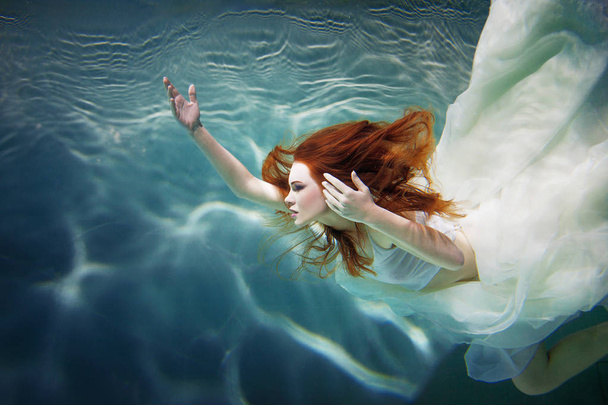 Underwater girl. Beautiful red-haired woman in a white dress, swimming under water. - Valokuva, kuva