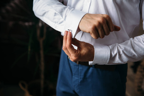 Close-up groom hands hold cufflinks. Elegant gentleman clother, white shirt and black belt. concept of classic work clothes in the office, jewelry. - Foto, Imagen