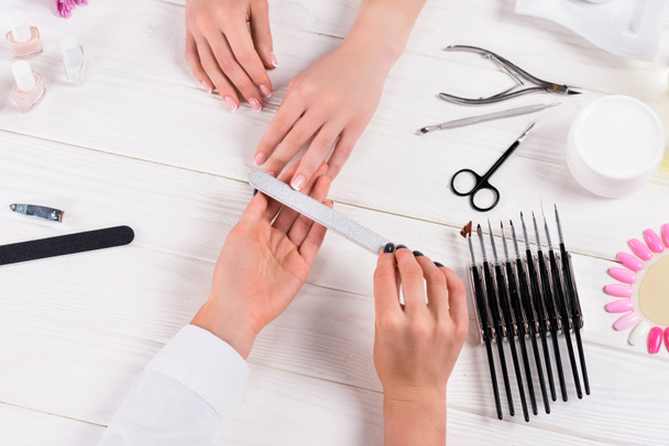 cropped image of beautician doing manicure by nail file to woman at table with cream, nail polishes, scissors, cuticle pusher, nail clippers, samples of nail varnishes  - Foto, Imagem