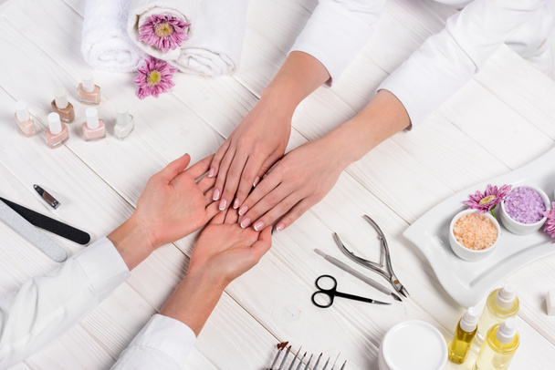 cropped image of manicurist looking at hands of woman at table with flowers, towels, nail polishes, nail files, nail clippers, sea salt, cream, cuticle pusher, scissors and aroma oil bottles - Photo, Image