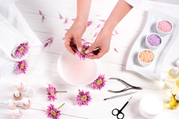 Μερική άποψη της γυναίκας που ρίχνει ροδοπέταλα στο λουτρό για νύχια στο τραπέζι με λουλούδια, πετσέτες, πολύχρωμο θαλασσινό αλάτι, άρωμα μπουκάλια λάδι, βερνίκια νυχιών, κρέμα εμπορευματοκιβωτίων και εργαλεία για μανικιούρ στο κομμωτήριο  - Φωτογραφία, εικόνα