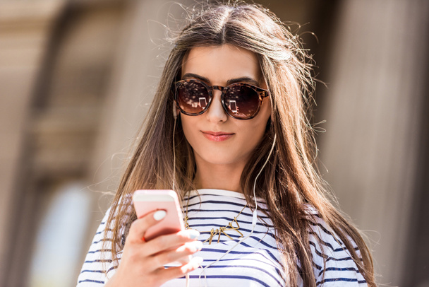 Porträt einer jungen attraktiven Frau mit Sonnenbrille mit Smartphone auf der Straße - Foto, Bild