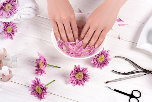 tiro cortado de mulher recebendo banho com pétalas para unhas à mesa com flores, toalhas, esmaltes e ferramentas para manicure no salão de beleza
  - Foto, Imagem