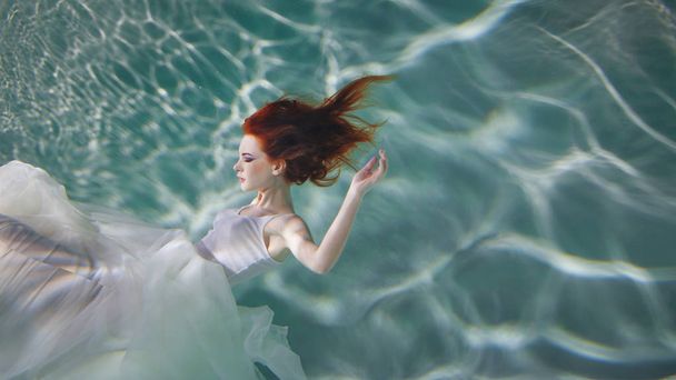 Underwater girl. Beautiful red-haired woman in a white dress, swimming under water. - Photo, Image
