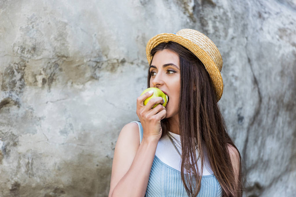 portrait of young stylish woman in hat eating fresh apple on street - Fotoğraf, Görsel