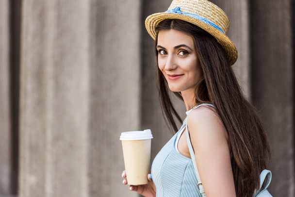 Seitenansicht der hübschen lächelnden Frau mit Strohhut und Kaffee zum Straßengehen  - Foto, Bild