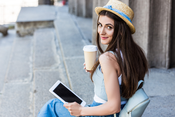 vista lateral de la joven mujer sonriente con la tableta y el café para ir sentado en los pasos
 - Foto, imagen