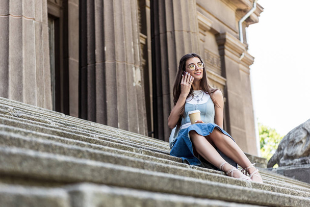 young smiling woman with coffee to go talking on smartphone while sitting on steps on street - Photo, image