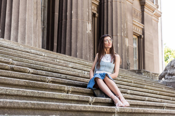 young stylish woman in eyeglasses and denim skirt resting on steps on street - Foto, Imagem