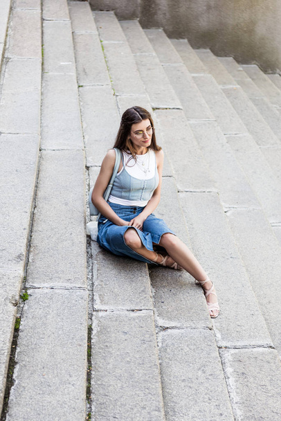 young stylish woman in eyeglasses and denim skirt resting on steps on street - Photo, Image