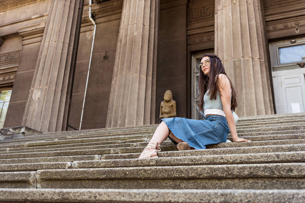junge stylische Frau mit Brille und Jeansrock ruht auf Stufen auf der Straße - Foto, Bild
