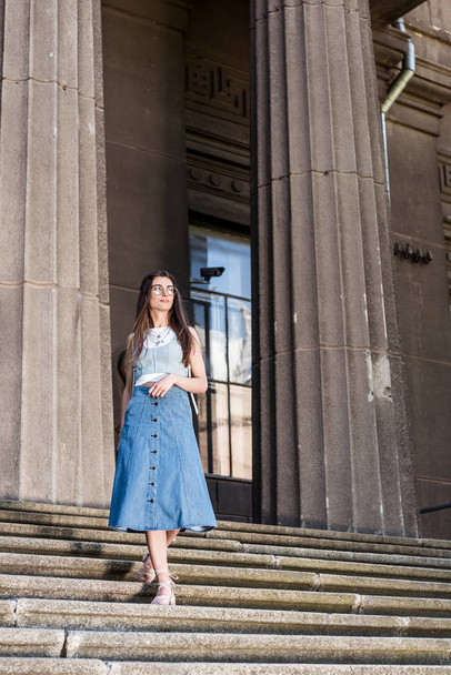 young stylish woman in eyeglasses and denim skirt standing on steps - Zdjęcie, obraz