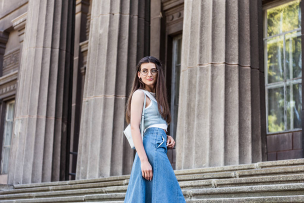 young stylish woman in eyeglasses and denim skirt standing on steps - Фото, зображення