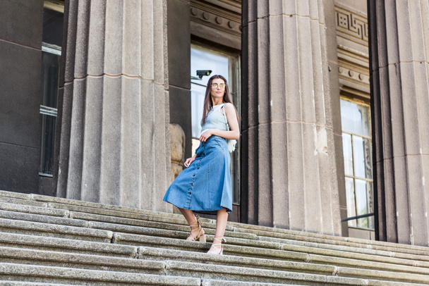 young stylish woman in eyeglasses and denim skirt standing on steps - Foto, imagen